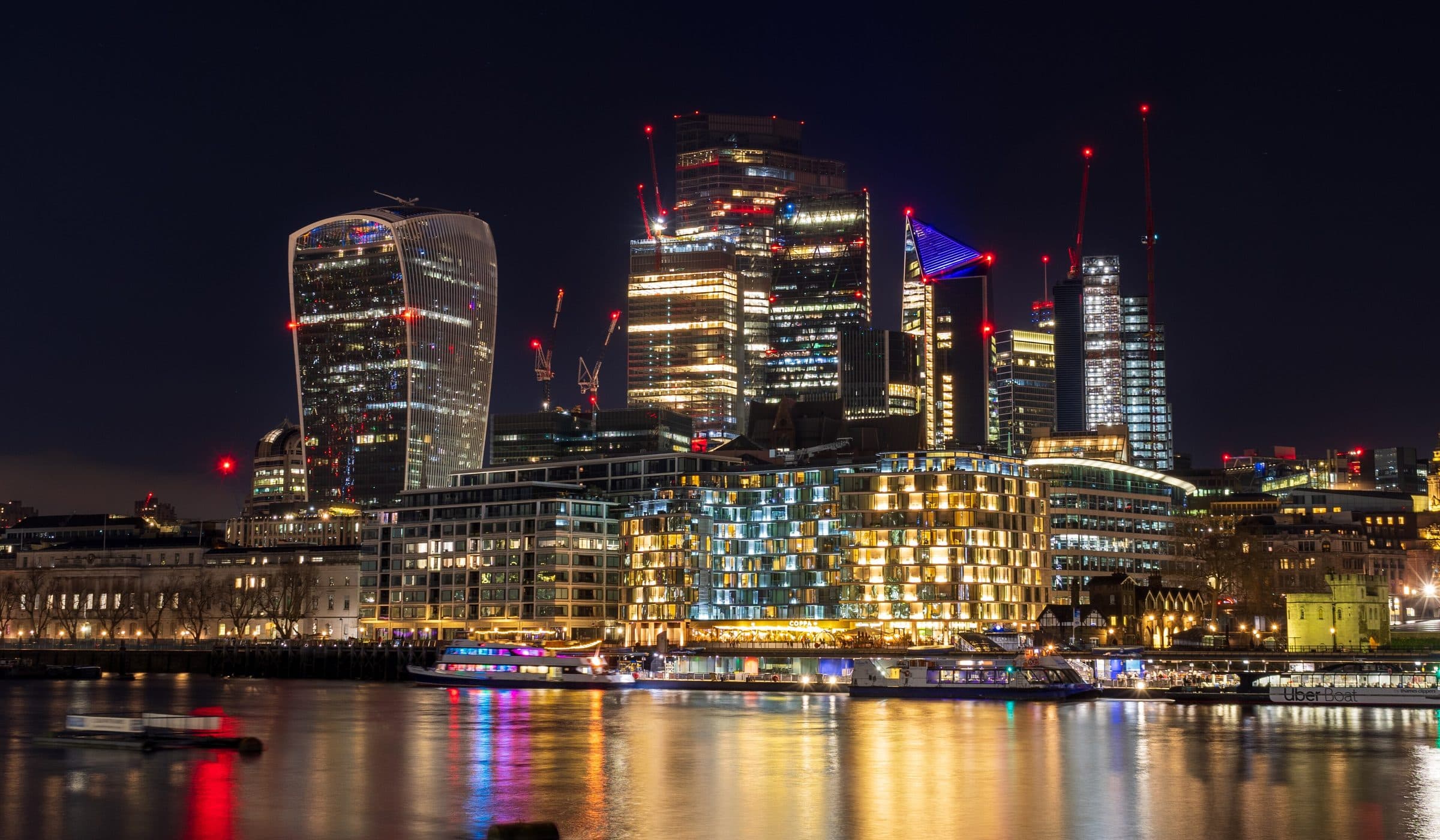 The City of London skyline at night, seen across the Thames, with the tower cluster lit and reflected in the water.